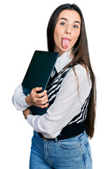 Young brunette teenager hugging laptop with love sticking tongue out happy with funny expression.