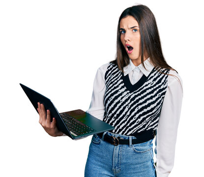 Young brunette teenager working using computer laptop scared and amazed with open mouth for surprise, disbelief face