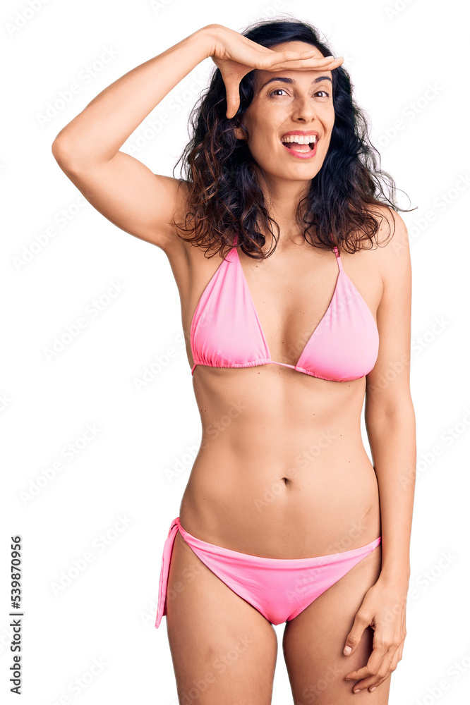 Wall mural Young beautiful hispanic woman wearing bikini very happy and smiling looking far away with hand over head. searching concept.