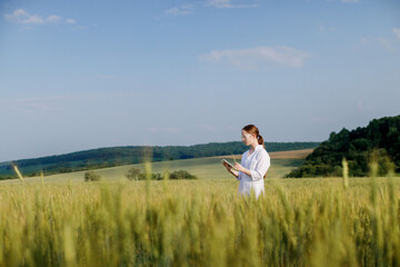 Naklejka na ściany i meble Female environmentalist scientist in a white coat in a green field of unripe ears of corn and recording data on a tablet device. Female researcher, biologist, walking in the field