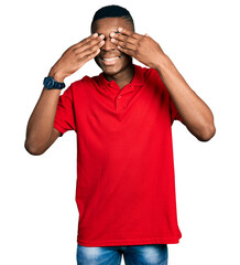 Young african american man wearing casual red t shirt covering eyes with hands smiling cheerful and funny. blind concept.