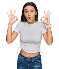 Young hispanic girl wearing casual white t shirt looking surprised and shocked doing ok approval symbol with fingers. crazy expression