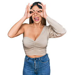 Young hispanic woman wearing casual clothes doing ok gesture like binoculars sticking tongue out, eyes looking through fingers. crazy expression.