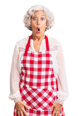 Senior grey-haired woman wearing apron scared and amazed with open mouth for surprise, disbelief...