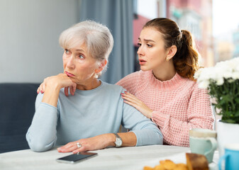 Depressed mature woman sitting at table and her adult daughter trying to encourage her.
