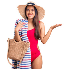 Young beautiful latin girl wearing swimwear and summer hat holding beach towel and bag celebrating victory with happy smile and winner expression with raised hands
