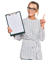 Beautiful blonde business woman holding clipboard with blank space surprised with an idea or...