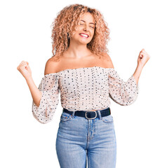 Young blonde woman with curly hair wearing casual clothes very happy and excited doing winner...
