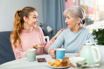 Friendly family of elderly mother and young adult daughter having good time while drinking tea and chatting happily in living room.