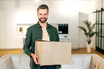 Male with cardboard box. Happy stylish caucasian man, stand at home in living room, holding a large cardboard box, received parcel from the online store, preparing to unpack, looks at camera, smile