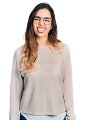 Beautiful hispanic woman wearing casual sweater and glasses sticking tongue out happy with funny expression. emotion concept.