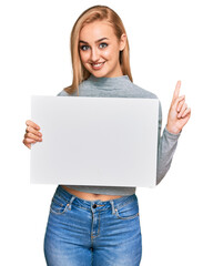 Fototapeta premium Beautiful caucasian woman holding blank empty banner surprised with an idea or question pointing finger with happy face, number one