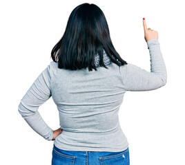 Young hispanic plus size woman wearing winter scarf posing backwards pointing ahead with finger hand