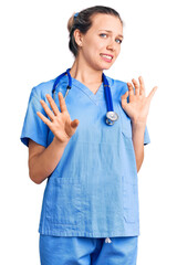 Young beautiful blonde woman wearing doctor uniform and stethoscope disgusted expression, displeased and fearful doing disgust face because aversion reaction. with hands raised