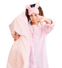 Beautiful kid girl with curly hair wearing sleep mask and pajamas holding pillow with angry face, negative sign showing dislike with thumbs down, rejection concept