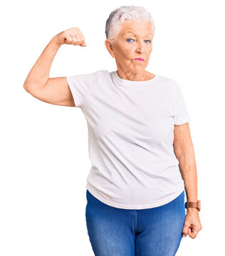 Senior beautiful woman with blue eyes and grey hair wearing casual white tshirt strong person showing arm muscle, confident and proud of power