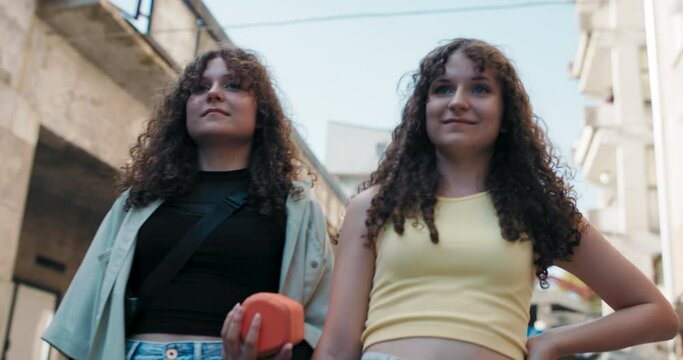Two teenage girls with beautiful curly hair walk through a residential area in the city holding a portable speaker to listen to music. The girls look at each other and forward.