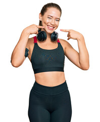 Beautiful blonde woman wearing gym clothes and using headphones smiling cheerful showing and pointing with fingers teeth and mouth. dental health concept.