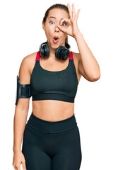 Beautiful blonde woman wearing gym clothes and using headphones doing ok gesture shocked with surprised face, eye looking through fingers. unbelieving expression.