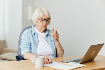 Fototapeta na wymiar an elderly woman is sitting at home in a bright interior and carefully looking at a laptop monitor, working with her index finger raised up, talking in an online conference with a colleague