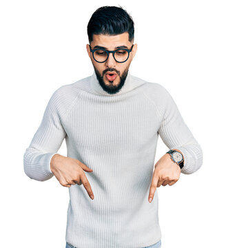 Young arab man with beard wearing elegant turtleneck sweater and glasses pointing down with fingers showing advertisement, surprised face and open mouth