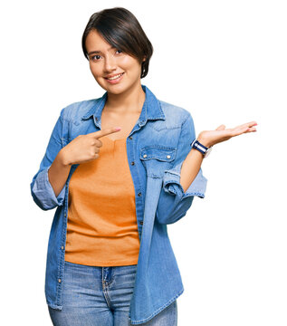 Young beautiful hispanic woman with short hair wearing casual denim jacket amazed and smiling to the camera while presenting with hand and pointing with finger.