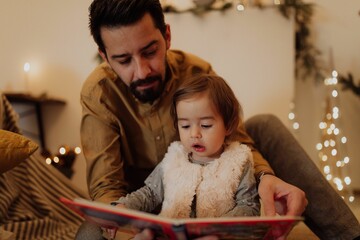 Father and his daughter reading a a book with Christmas decoration in the background. - obrazy, fototapety, plakaty