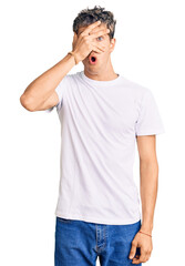 Young handsome man wearing casual white tshirt peeking in shock covering face and eyes with hand, looking through fingers with embarrassed expression.