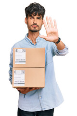 Young hispanic man holding delivery package with open hand doing stop sign with serious and confident expression, defense gesture