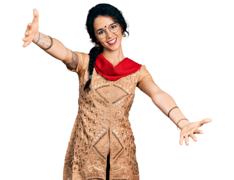 Young indian woman wearing traditional dress and glasses looking at the camera smiling with open arms for hug. cheerful expression embracing happiness.