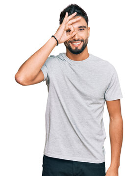 Young man with beard wearing casual grey tshirt doing ok gesture with hand smiling, eye looking through fingers with happy face.