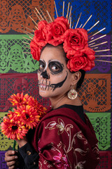 Mexican woman in traditional make up and Catrina costume, holding an orange rose. Colorful cut paper background