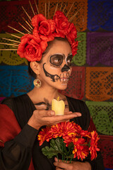 Mexican woman in traditional make up and Catrina costume, holding a candle and a bouquet of orange flowers. Colorful cut paper background