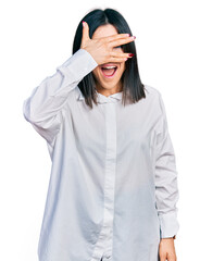 Young brunette woman with blue eyes wearing oversize white shirt peeking in shock covering face and eyes with hand, looking through fingers with embarrassed expression.