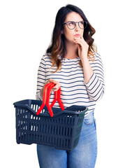 Beautiful young brunette woman holding supermarket shopping basket serious face thinking about question with hand on chin, thoughtful about confusing idea