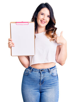 Beautiful young brunette woman holding clipboard with blank space pointing finger to one self smiling happy and proud