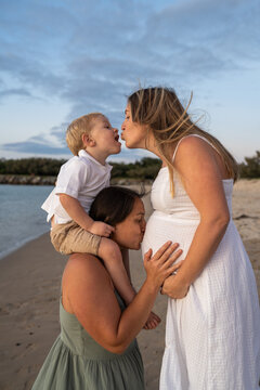 toddler on shoulders of lesbian mother kissing other mom at the beach