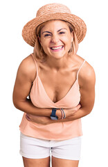 Young blonde woman wearing summer hat smiling and laughing hard out loud because funny crazy joke with hands on body.