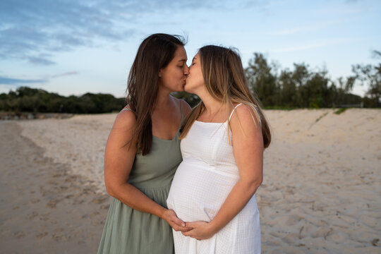 lesbian couple kissing on the beach holding pregnant belly