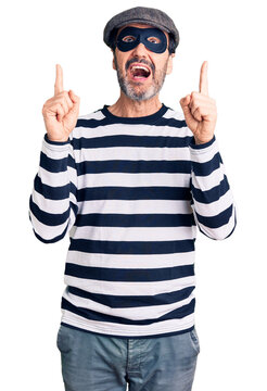 Middle age handsome man wearing burglar mask amazed and surprised looking up and pointing with fingers and raised arms.