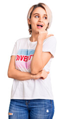 Young beautiful blonde woman wearing t shirt with diversity word message smiling with happy face looking and pointing to the side with thumb up.