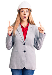 Beautiful young woman wearing architect hardhat amazed and surprised looking up and pointing with fingers and raised arms.