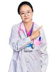 Young chinese woman wearing doctor uniform and stethoscope pointing with hand finger to the side showing advertisement, serious and calm face