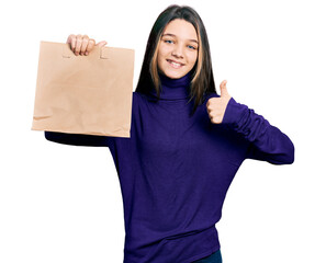 Young brunette girl with long hair holding take away paper bag smiling happy and positive, thumb up doing excellent and approval sign