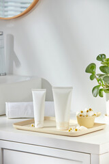 Cosmetics sample on a beige wooden tray with many white flowers, green plants, and towels on a white table inside ceramic bathroom background.