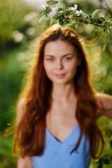 Beautiful woman with long red hair near a tree in the summer sun in the nature in the park smiling without allergies in a blue dress, the concept of health and beauty
