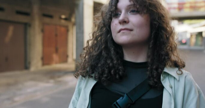 Close-up of the face of a young teenage girl with curly hair standing by the garages in a residential area holding an orange portable speaker to listen to music.