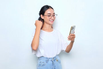 Excited young Asian girl in white t-shirt using smartphone and doing winner gesture isolated on...