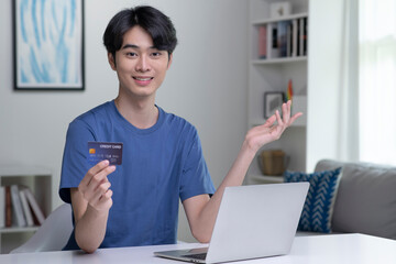 Portrait of young Asian man looking at camera while holding credit card and using laptop computer....