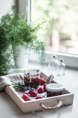 Jars with strawberry jam, strawberries on a linen towel and a white wooden tray and flowers in a vase on the windowsill.
